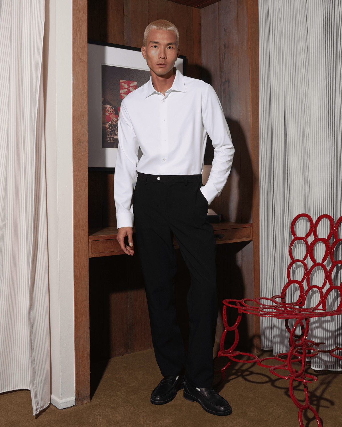 A person standing in a room with white curtains and a textured wall, wearing a white classic non-iron shirt with a refined collar, paired with dark trousers and black shoes.